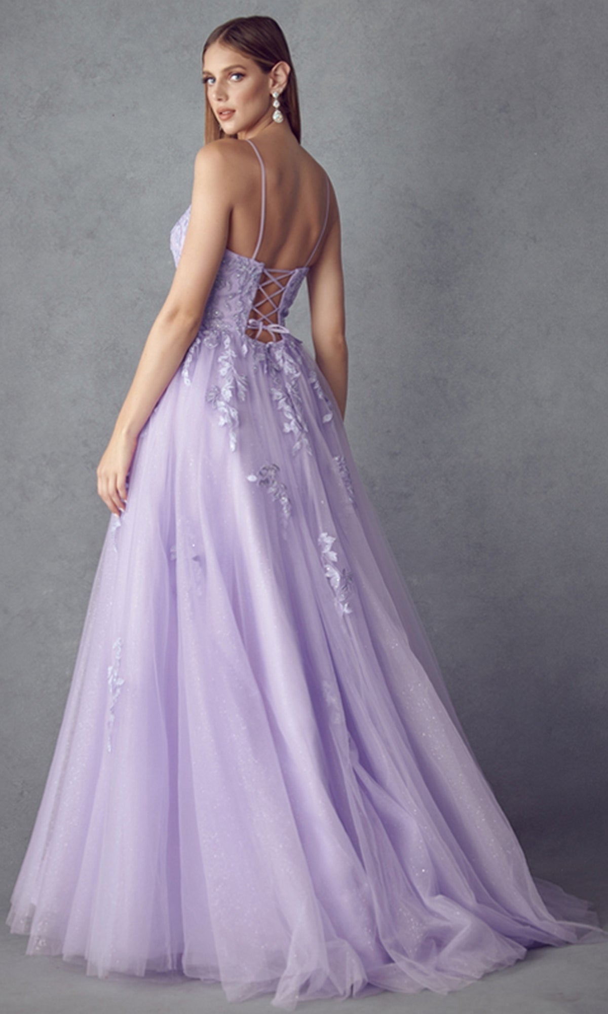 A woman in the Corset-Back Long Glitter-Tulle Prom Ball Gown 260 stands against a gray backdrop, looking over her shoulder.